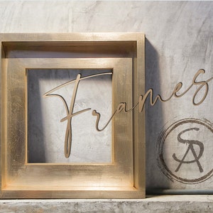 Könnte beinhalten: Ein goldfarbener Bilderrahmen mit einem Doppelrahmen-Design. Der Rahmen steht auf einer grauen, strukturierten Oberfläche. Das Wort "Rahmen" ist in Kursivschrift über dem Rahmen geschrieben.