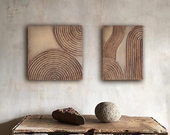 Op de afbeelding: Twee vierkante wandkunstwerken met een beige achtergrond en bruine, concentrische halve cirkelontwerpen. Daaronder toont een verweerde houten tafel een donkere, onregelmatig gevormde steen en een ronde, grijze steen.