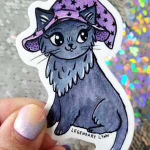 May include: A sticker of a cartoon cat wearing a purple witch's hat decorated with black stars. The cat is depicted in shades of grey with large eyes and a fluffy chest. The sticker has the text "LEGENDARY LYNN".