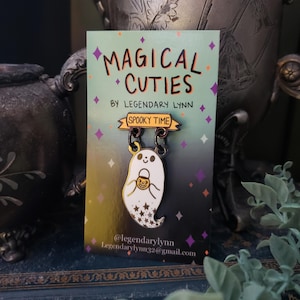 May include: A pair of enamel pins featuring a ghost holding a candy bucket and a banner that reads "Spooky Time". The pins are on a card with the text "Magical Cuties by Legendary Lynn" and the social media handle "@legendarylynn".