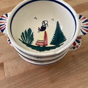 May include: Stacked ceramic bowls with a cream-colored interior and a blue rim. Each bowl features a hand-painted design of a woman in traditional clothing, holding flowers, with green foliage and a tree. The handles are decorated with red, white, and blue accents.