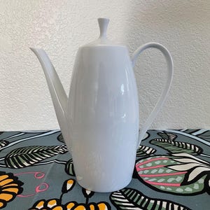 Puede incluir: Una cafetera de cerámica blanca con un cuerpo alto y delgado y una tapa redonda. La cafetera tiene un pico en un lado y un asa en el otro.