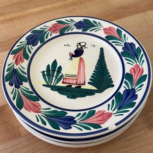 May include: A stack of three ceramic plates with a hand-painted design. Each plate features a central image of a figure in traditional clothing, surrounded by a border of blue, green, and pink floral motifs. The plates have a white background and a blue rim.