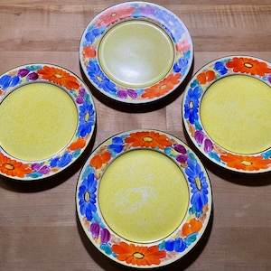 May include: Four yellow ceramic plates with a floral border. The border features a pattern of blue, orange, purple, and pink flowers. The plates have a black rim and are arranged on a wooden surface.