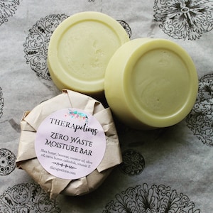 Peut inclure: Deux barres de soin hydratantes zéro déchet rondes de couleur jaune pâle enveloppées dans du papier brun avec une étiquette blanche qui indique "THERApotions ZERO WASTE MOISTURE BAR Shea butter, beeswax, coconut oil, olive oil, cocoa butter, calendula, vitamin E. therapotions.com"