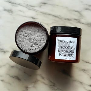 Peut inclure: Deux pots en verre avec des couvercles noirs. Un pot contient une poudre grise et l'autre pot porte une étiquette qui indique "THERApeltions TOOTH BRUSHING POWDER 4 oz."