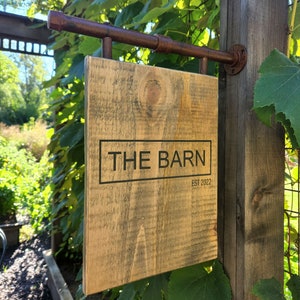 May include: A rustic wooden sign with the text "THE BARN EST 2022" hanging from a rusty metal pipe. The sign is mounted on a wooden post and surrounded by green foliage.