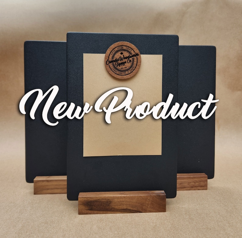 Puede incluir: Tres letreros negros de pizarra con bases de madera. El letrero del medio tiene un c&iacute;rculo de madera marr&oacute;n con el texto "Canada Woodworks Signage Co." en &eacute;l. El texto "New Product" est&aacute; escrito en cursiva blanca en los letreros.