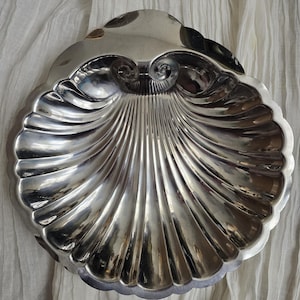 May include: A shiny, silver-colored seashell-shaped serving dish. The dish has a scalloped edge and a detailed, ribbed interior. The design includes two curled accents at the top. The dish is likely made of metal and reflects light.