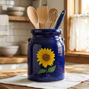 May include: A deep blue ceramic kitchen utensil crock featuring a vibrant yellow sunflower illustration with green leaves on the front. The cylindrical container holds several wooden spoons, a metal whisk, and a blue-handled kitchen tool.