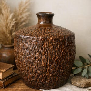 May include: A brown ceramic vase with a textured surface, reminiscent of tree bark. The vase has a rounded body and a narrow neck. It sits on a wooden surface, with other decorative items in the background.
