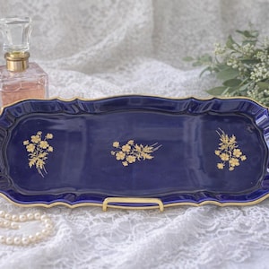 May include: A rectangular, navy blue ceramic tray with gold trim and floral accents. The tray has a scalloped edge and gold handles. A perfume bottle and a strand of pearls are next to the tray.