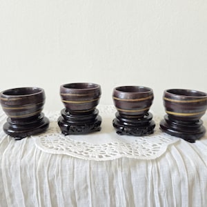 May include: Four small, dark brown ceramic bowls with a yellow stripe, each resting on a dark wooden stand. The bowls have a slightly angled rim and are displayed on a white, textured surface.