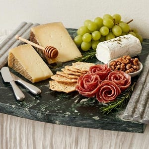 May include: A rectangular dark green marble serving board with fluted gray stone handles. The board is arranged with two wedges of hard cheese, a log of goat cheese, green grapes, crackers, salami roses, walnuts, a honey dipper, and a metal knife.