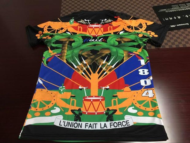 May include: Black t-shirt with a colorful design featuring a coat of arms with palm trees, cannons, drums, and the text "L'UNION FAIT LA FORCE".