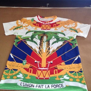 May include: White t-shirt with a colorful print of the Haitian flag. The flag features a palm tree, cannons, and a drum. The text "L'UNION FAIT LA FORCE" is printed at the bottom of the shirt.