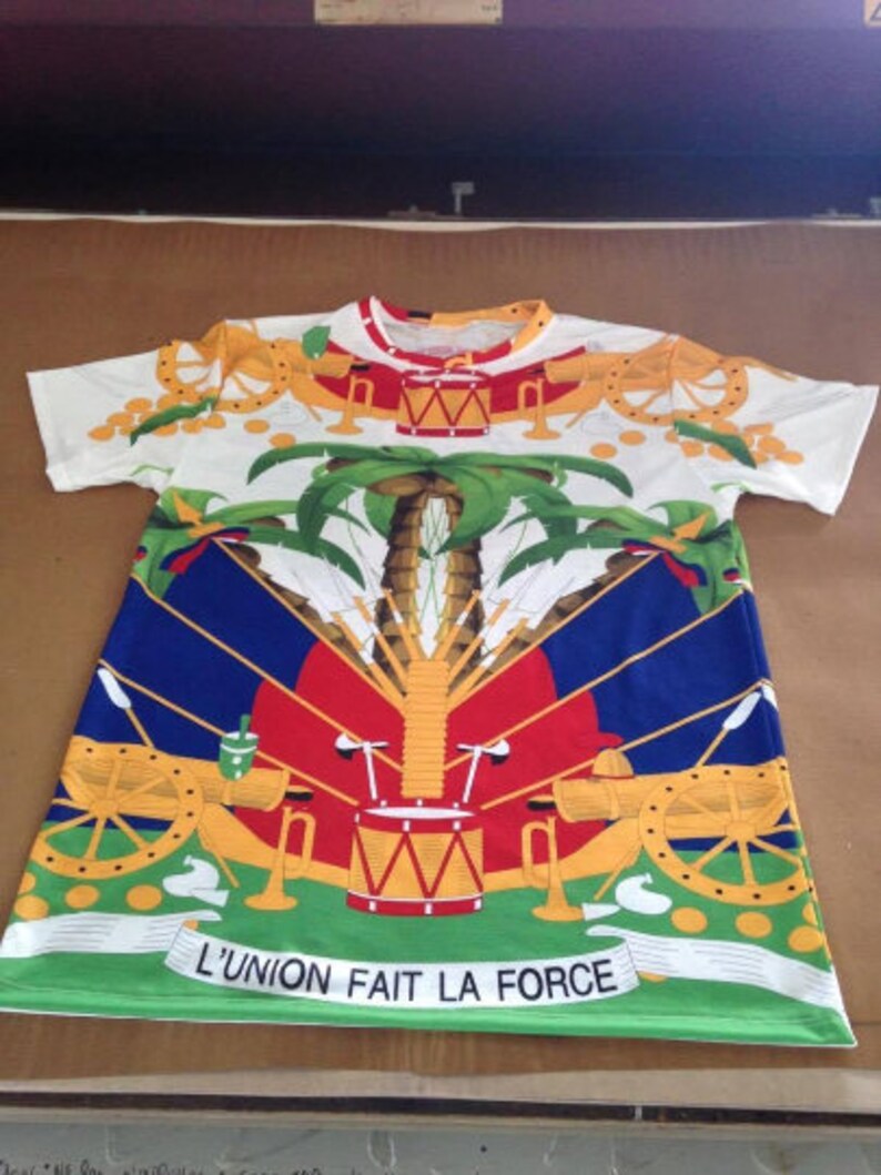 May include: White t-shirt with a colorful print of the Haitian flag. The flag features a palm tree, cannons, and a drum. The text "L'UNION FAIT LA FORCE" is printed at the bottom of the shirt.