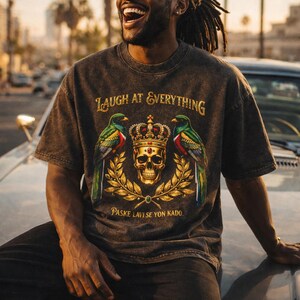 Puede incluir: Camiseta gris oscuro con el texto "LAUGH AT EVERYTHING" sobre una calavera coronada, flanqueada por dos pájaros. Debajo está el texto "PASKE LAVI SE YON KADO."