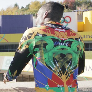 May include: A person wearing a long-sleeved shirt with a colorful, abstract design. The shirt features a green palm tree, a red and yellow drum, and the text "Ha&iuml;ti" in blue. The shirt also has the text "L'UNION FAIT LA FORCE" in white.