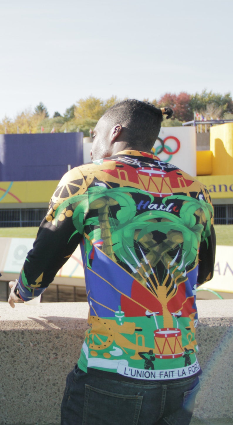 May include: A person wearing a long-sleeved shirt with a colorful, abstract design. The shirt features a green palm tree, a red and yellow drum, and the text "Ha&iuml;ti" in blue. The shirt also has the text "L'UNION FAIT LA FORCE" in white.