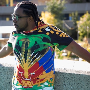 May include: A man wearing a black, green, yellow, red, and blue t-shirt with a palm tree and a coat of arms design. The shirt has a  "UNITE" text on the bottom.