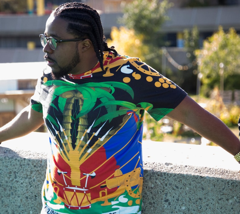 May include: A man wearing a black, green, yellow, red, and blue t-shirt with a palm tree and a coat of arms design. The shirt has a  "UNITE" text on the bottom.