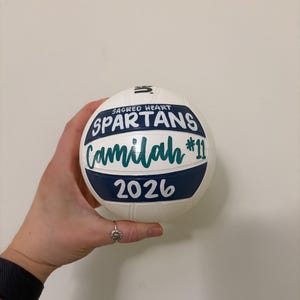 May include: A white volleyball featuring dark blue panels with the text SACRED HEART SPARTANS in white, the name Camilah in teal script, the number 11 in white, and the year 2026 in white. The ball has a smooth surface and visible seams.