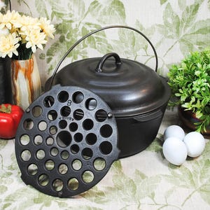 May include: A black cast iron Dutch oven with a lid and handle, a trivet, and a red tomato. The Dutch oven is next to a small potted plant and three white eggs. A vase of yellow flowers and a metal bucket are in the background.