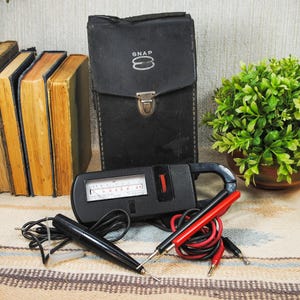 May include: A black clamp-style multimeter with an analog display, featuring a red thumbwheel and a set of black and red test leads. The device is accompanied by a black carrying case with the word SNAP printed in white on the front flap.