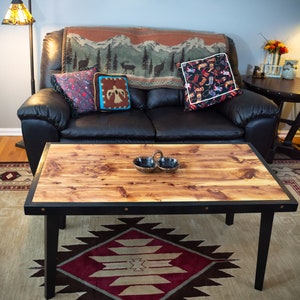 May include: A rustic wooden coffee table with a black metal frame and a natural wood top. The table is sitting on a rug with a geometric pattern. There are two small bowls on the table.