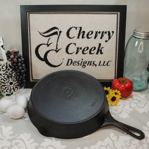 May include: Black cast iron skillet with a raised logo that reads "The Original Lodge Cast Iron", a wooden handle, and a white background.