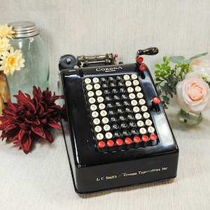 May include: A vintage black Corona typewriter with white number keys and red function keys. The typewriter is positioned next to a vase of yellow flowers, a jar, and a bouquet of pink roses. The typewriter has the text "Corona" on it.