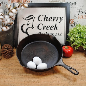 May include: A black cast iron skillet with three white eggs inside. The skillet is on a wooden surface, with a framed sign that reads "Cherry Creek Designs, LLC" in the background. Decorative elements include cotton stems, pine cones, and a red tomato.