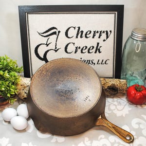 May include: A vintage cast iron skillet with a brass handle. The skillet is dark brown with signs of use. The image also includes a framed sign that reads "Cherry Creek Designs, LLC", a glass jar, a red tomato, and three white eggs.