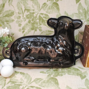 May include: A black lamb-shaped baking mold with handles. The mold is made of metal and has a glossy finish. Three white eggs and a bouquet of flowers are in the foreground. Vintage books are in the background.