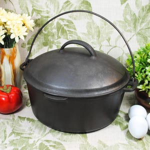May include: A black cast iron Dutch oven with a lid and a handle. The pot is round and sits on a floral patterned surface. A red tomato, white eggs, and yellow flowers are in the background.
