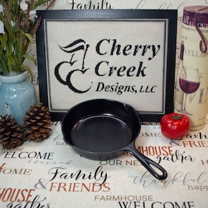 May include: A black cast iron skillet with a wooden handle sits on a fabric background with the words "Family & Friends" printed on it. The skillet is next to a framed cross-stitch that reads "Cherry Creek Designs, LLC".