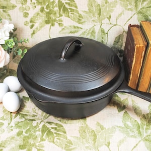May include: A black cast iron skillet with a lid, featuring a handle and a rounded shape. The skillet is displayed on a floral patterned surface, with white eggs and flowers in the background. The skillet's diameter is approximately 10 inches.