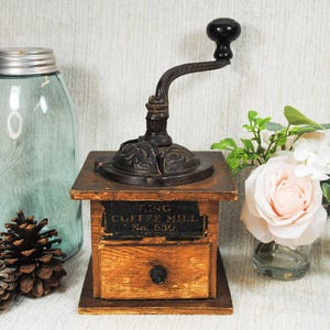 May include: An antique King Coffee Mill No. 630 with a wooden base and a metal grinder. A blue glass jar, pine cones, and a small bouquet of flowers are also in the image.