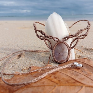May include: A brown macrame necklace with a light brown oval gemstone pendant. The necklace is lying on a brown leaf and sand.