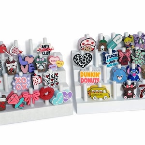 May include: A collection of colorful shoe charms displayed on white tiered stands. Charms include hearts, bows, cartoon characters, and phrases like "LOVE" and "Dunkin' Donuts". The charms are made of rubber and have a variety of colors.