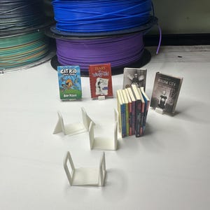 May include: An assortment of miniature books, including "Cat Kid Comic Club" and "Diary of a Wimpy Kid," are displayed with white book stands. Spools of blue, purple, and black filament are visible in the background, suggesting a 3D printing theme.
