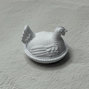 May include: A white, 3D-printed hen figurine, sitting in a nest-like base. The hen has detailed feather and comb textures. The figurine is a decorative item, possibly for home decor or a collector's piece.