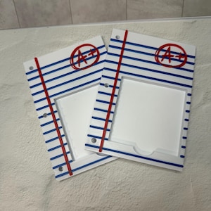 May include: Two white, notebook-style photo frames with blue lines and a red margin line. Each frame has a red circle with "A+" in the upper right corner. The frames are on a textured surface.