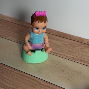 May include: A baby doll with blue eyes, brown hair, and a pink bow sits on a mint green potty. The doll wears a blue top and purple diaper. The doll is a miniature toy.