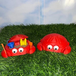 May include: Two red crab-shaped sand molds with white eyes and smiling faces sit on green grass. One crab has a hinged top, revealing a miniature sandcastle, bucket, rake, and shovel. The other crab is closed.