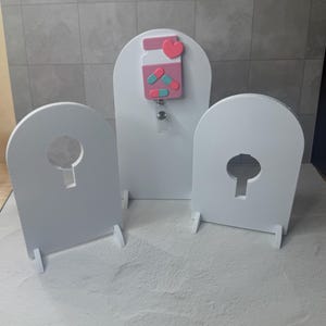 May include: Three white, arched display stands with keyhole cutouts. A badge reel with a pink pill bottle design, pink heart, and blue and pink pills is attached to the center stand. The stands are on a textured surface.