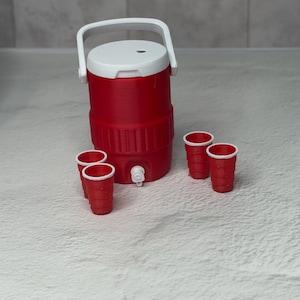 May include: A red and white miniature cooler with a white handle and a spigot, accompanied by six small red cups. The cooler has a cylindrical shape and a white lid. The cups are arranged around the cooler.