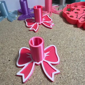 May include: A bright pink pen holder featuring a flat, bow-shaped base with a white outline and a cylindrical center tube. The object is constructed from layered material with visible texture lines, designed to stand upright on a flat surface.