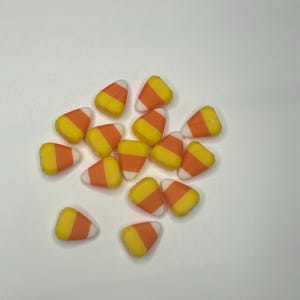 May include: A pile of candy corn pieces. Each piece has a white base, an orange center, and a yellow tip. The candies are scattered on a white surface.